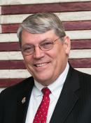 Image of Dennis Weber wearing a black suit and red tie, standing in front of a flag. Dennis is smiling and wearing glasses.