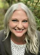 Jamie Merly smiling brightly at the camera with her long curly blonde hair, a white polka dot shirt and dark gray blazer. 