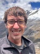 Danny wearing brown framed glasses, smiling at the camera with a large mountain scape behind him